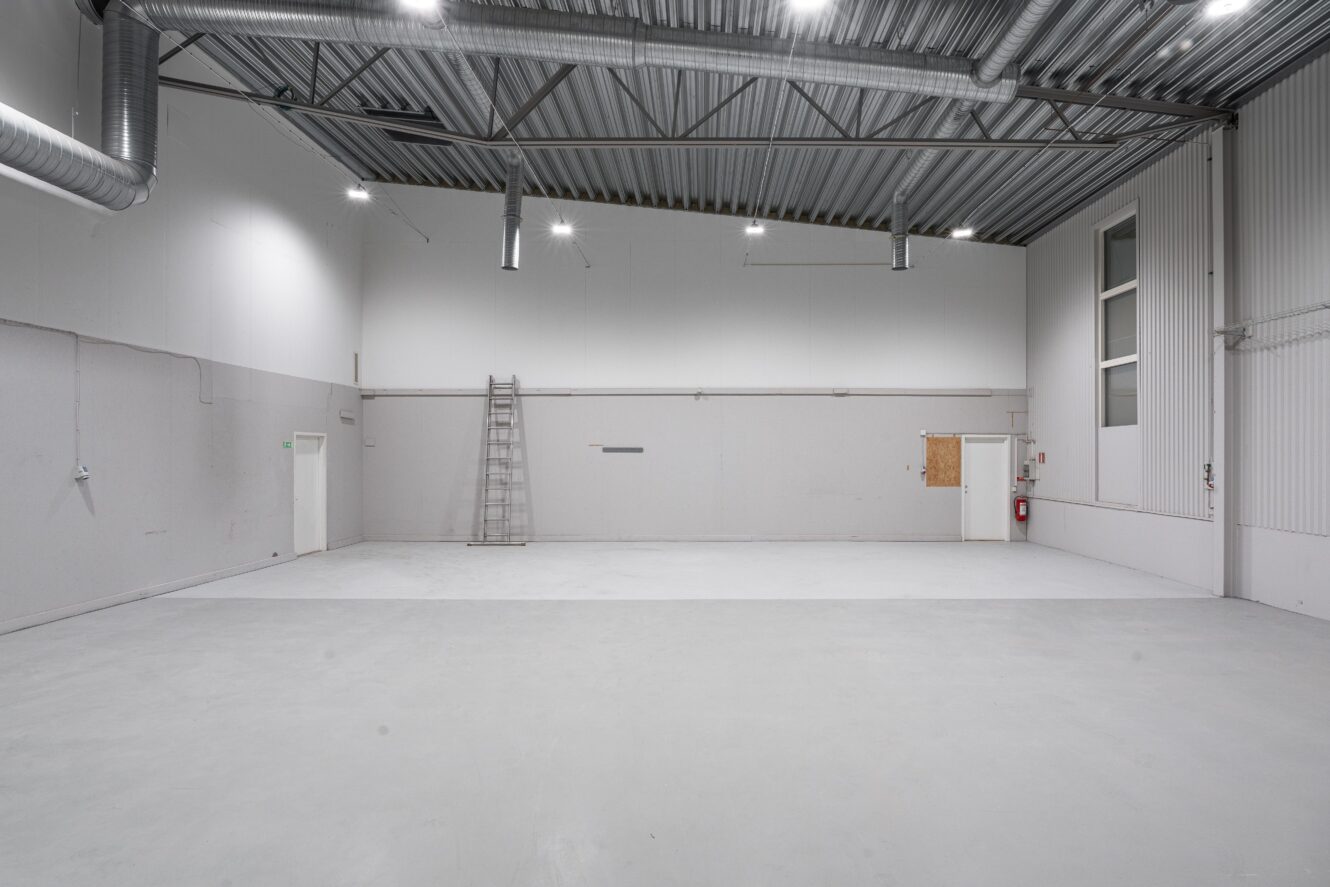 Tomt industriellt lagerutrymme med vita väggar, grått betonggolv, synliga rör i taket, en metallstege mot väggen och minimal belysning.