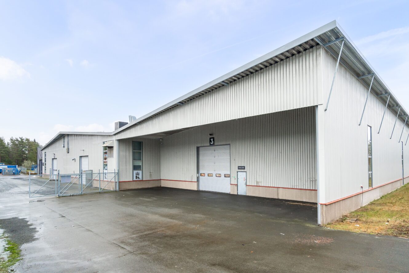 Ett stort, grått industrilager med korrugerad metallfasad, lastkaj, numrerad dörr och inhägnad entré en mulen dag.