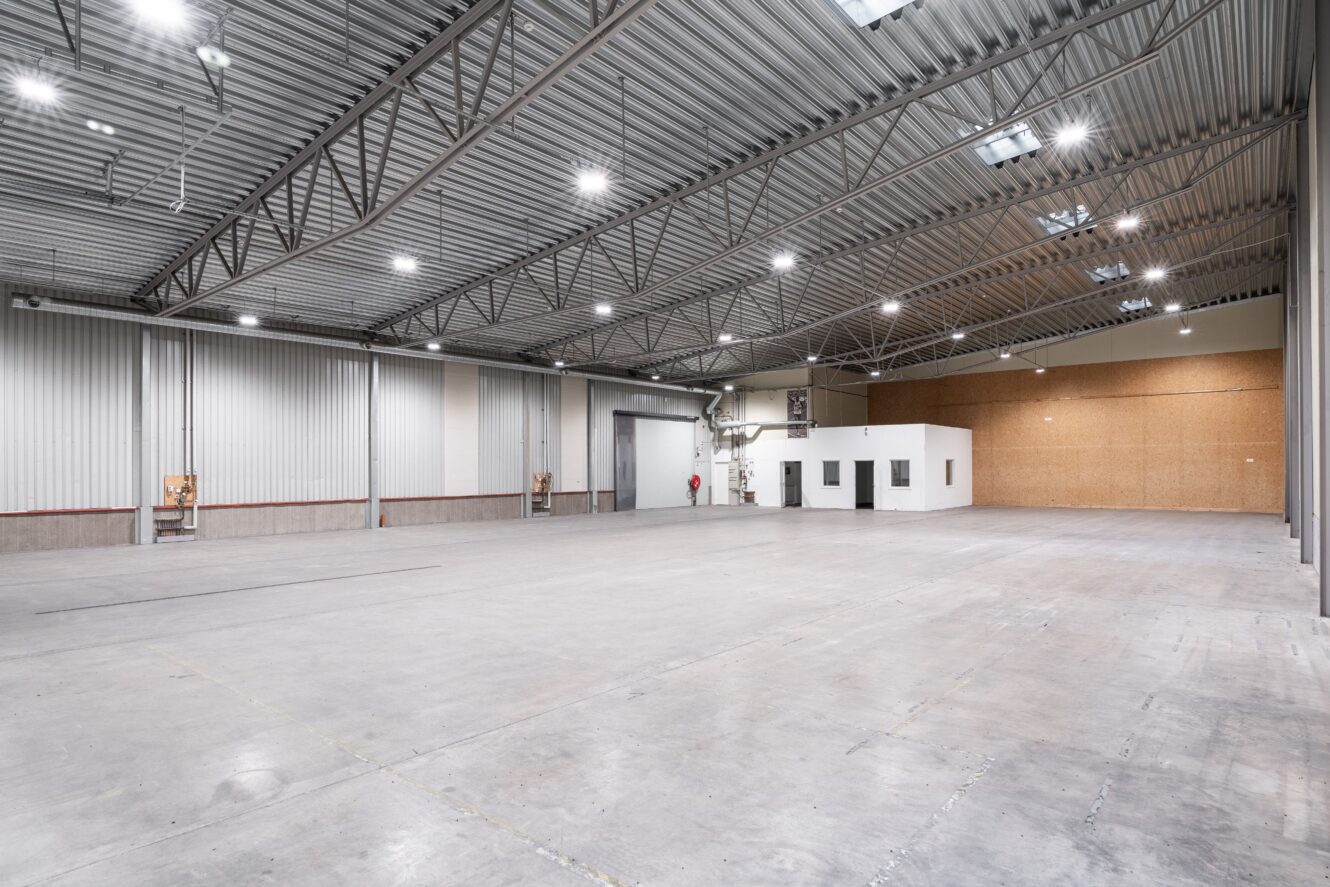 Stor, tom lagerlokal med högt metalltak, betonggolv, industriell belysning och en liten vit kontorsbyggnad i hörnet.