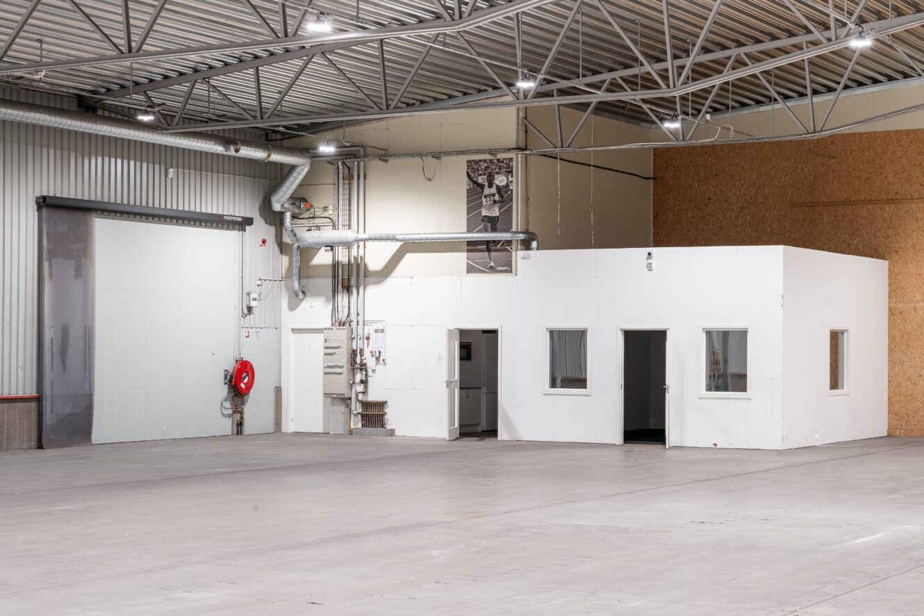 En vit, lådformad kontorsbyggnad med fönster ligger i ett rymligt, tomt industrilager med synliga takbjälkar och betonggolv.