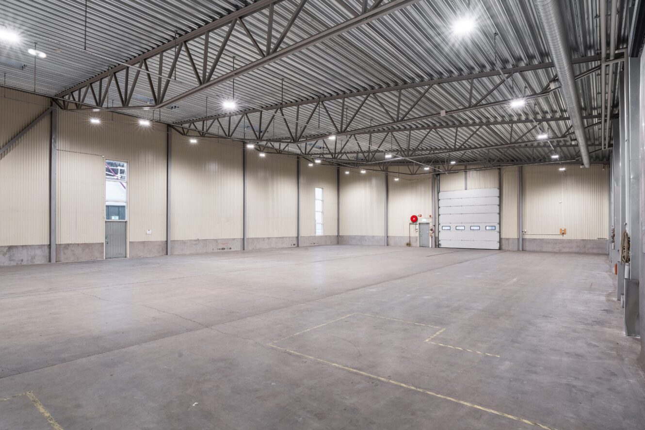 Stort, tomt industrilager med högt i tak, betonggolv, metallbalkar och takbelysning; en stor garageport och flera fönster syns.