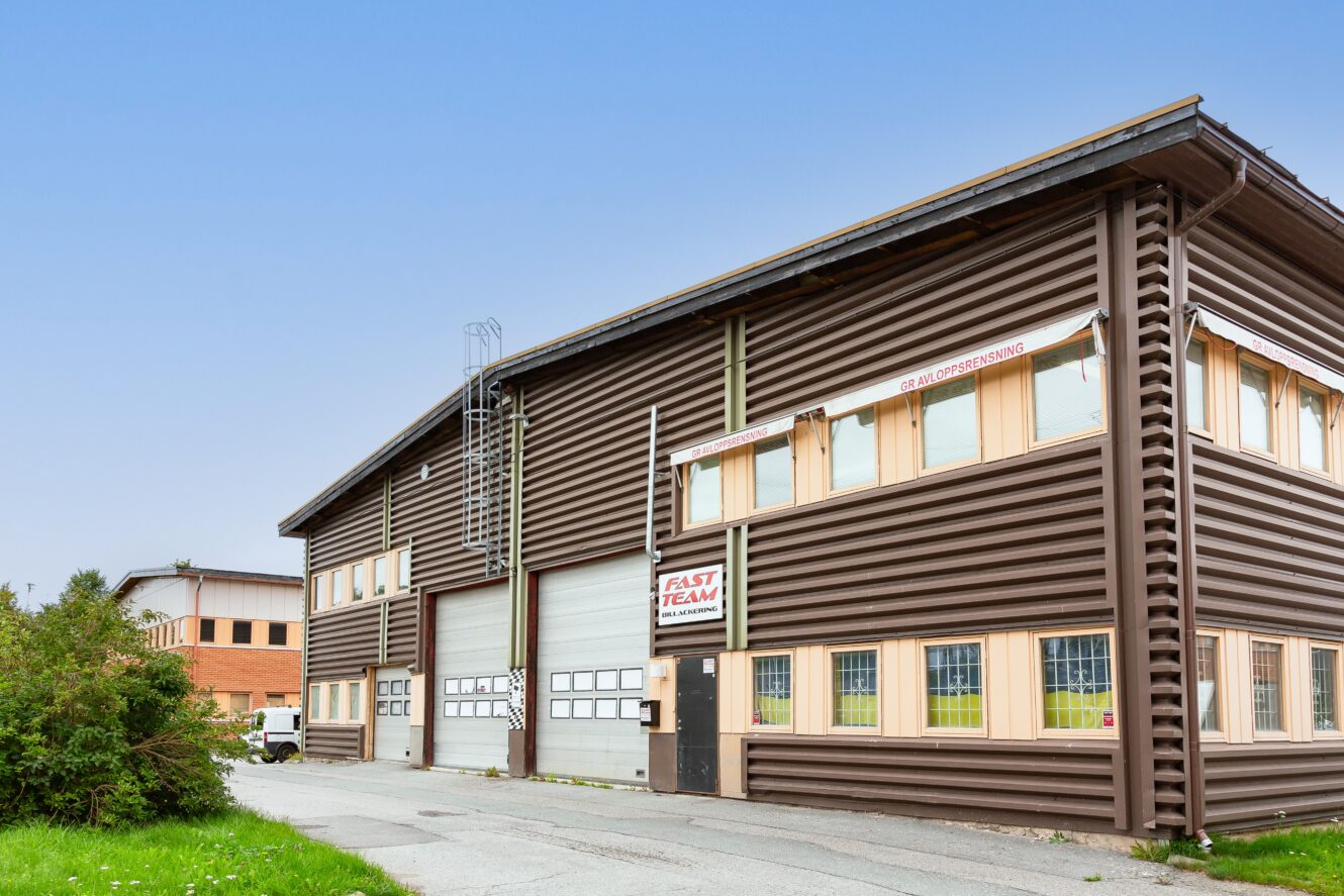 En stor industribyggnad i två våningar med timmerliknande fasad, flera garageportar och en skylt med texten FAST TEAM vid ingången.