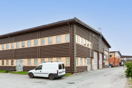 Industribyggnad i två våningar med brun fasadbeklädnad och beige fönsterkarmar, en vit skåpbil parkerad utanför och en klar himmel i bakgrunden.
