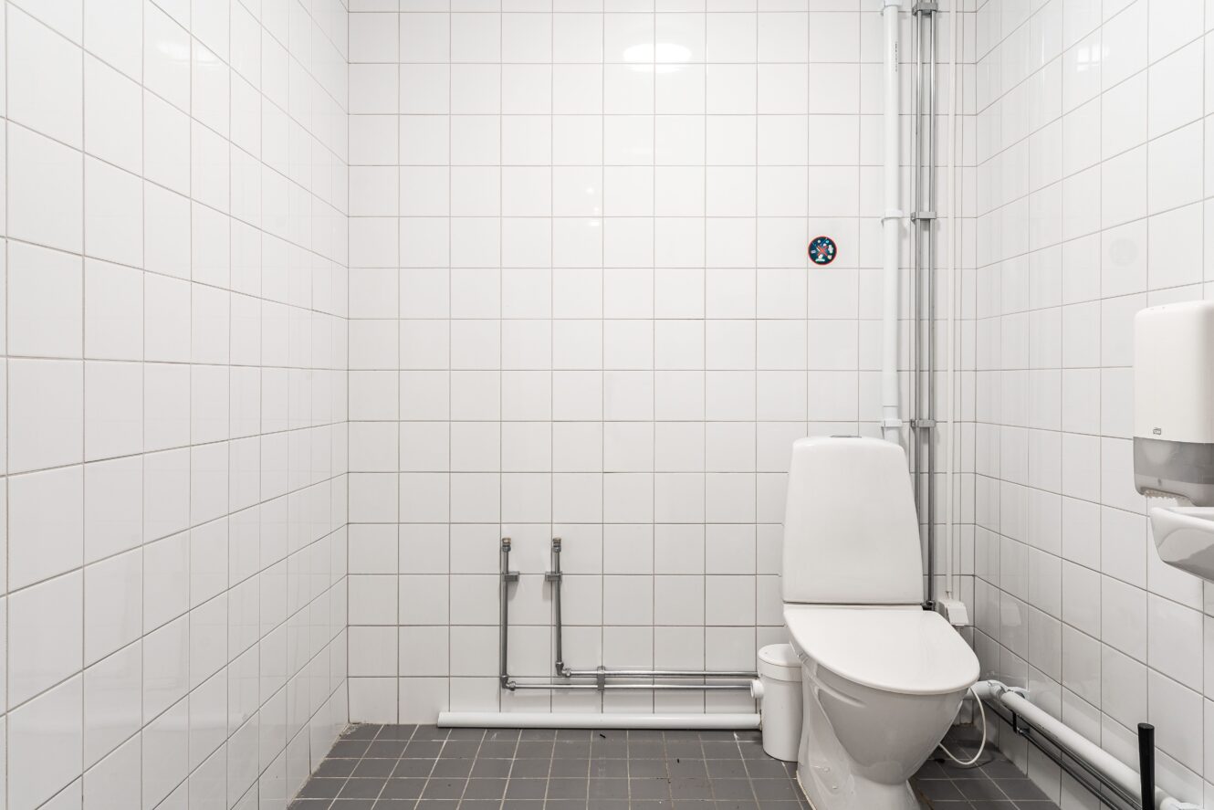 En vitkaklad toalett med en toalett, synliga rör, en väggmonterad toalettpappersbehållare och en liten skylt på väggen.