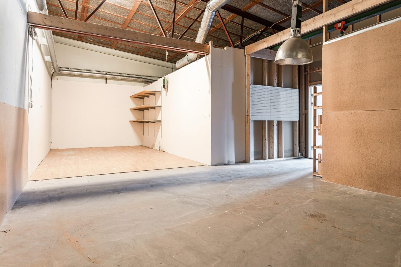 Interiör i en tom lagerlokal med betonggolv, synligt tak, en liten skiljevägg och trähyllor på en bakre vägg.