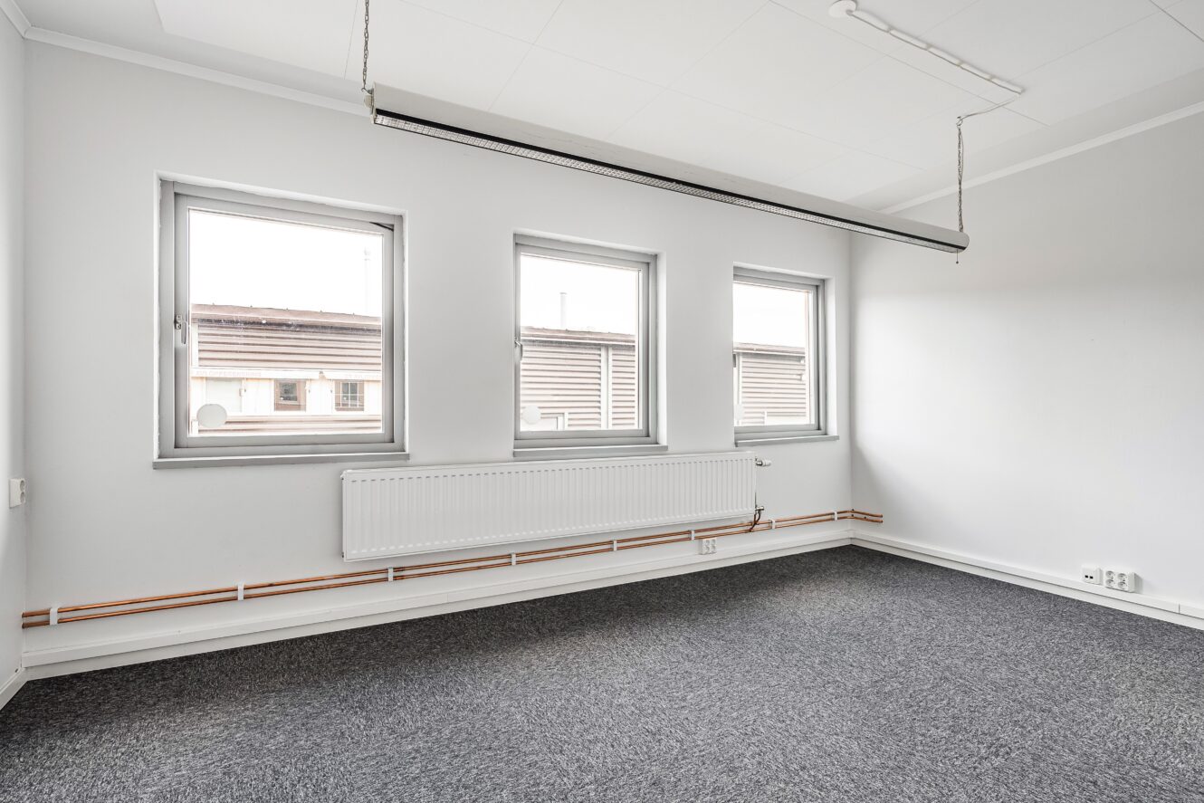 Ett tomt kontorsrum har tre fönster, vita väggar, en grå matta, ett element och lysrör i taket.
