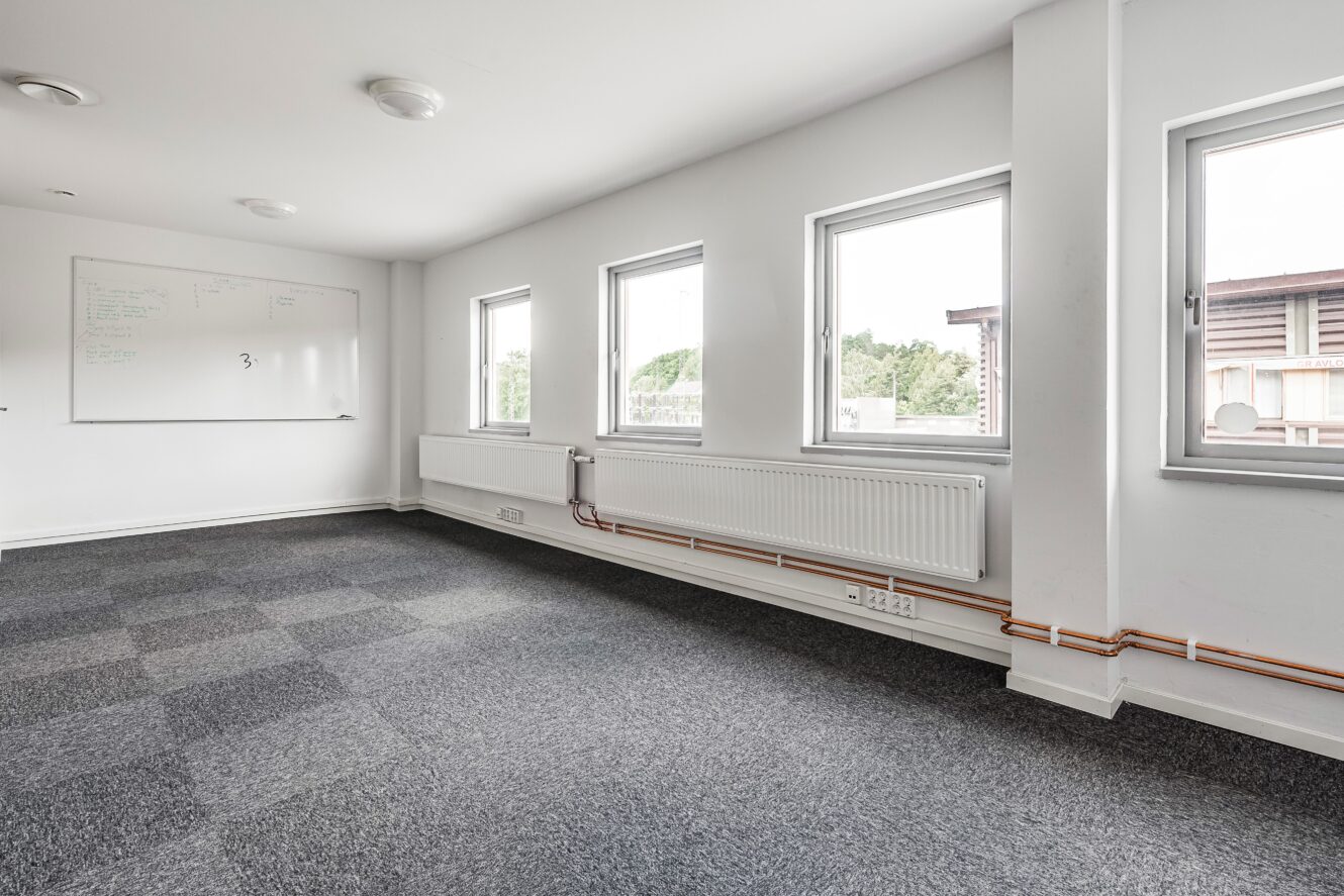 Ett tomt kontorsrum med grå heltäckningsmatta, tre stora fönster, en vit radiator, synliga rör och en whiteboard på väggen.