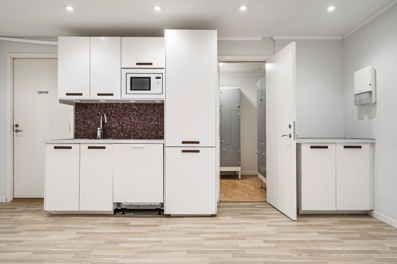 Ett litet, modernt pentry med vita skåp, mosaikplattor, inbyggd mikrovågsugn och en öppen dörr som leder till en tvättstuga med synliga maskiner.