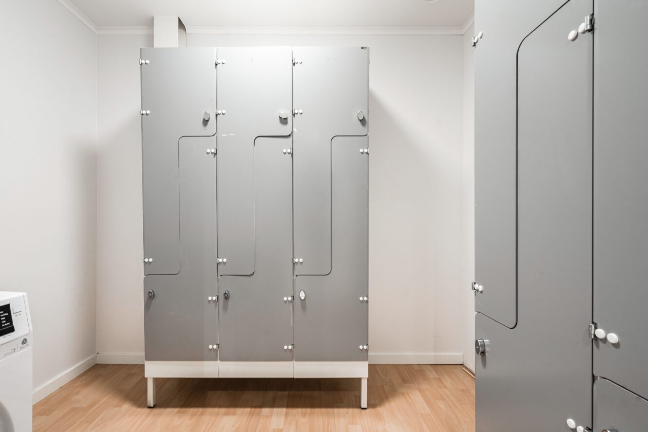 Ett rum med tre stängda grå metallskåp på ett ljust trägolv, vita väggar och en del av en vit tvättmaskin synlig till vänster.