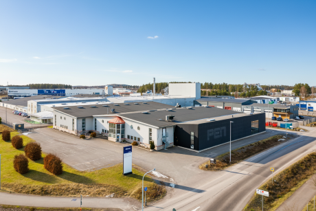 En stor industribyggnad med en svart sektion märkt PEN, omgiven av parkeringsplatser, vägar och skyltar, belägen i en företagspark en klar dag.
