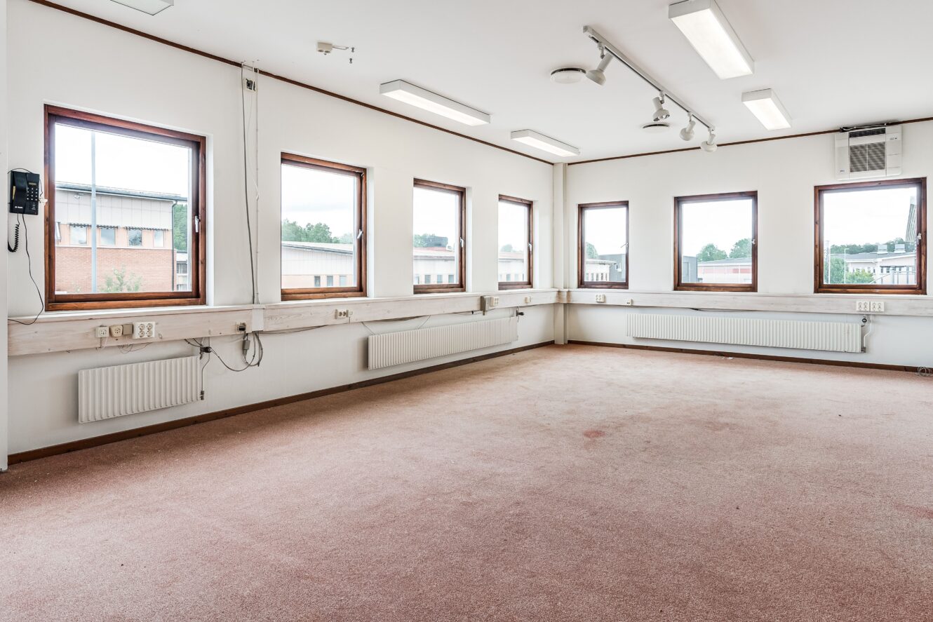 Tomt kontorsrum med beige matta, vita väggar, flera fönster, lysrör i taket, radiatorer och eluttag längs väggarna.