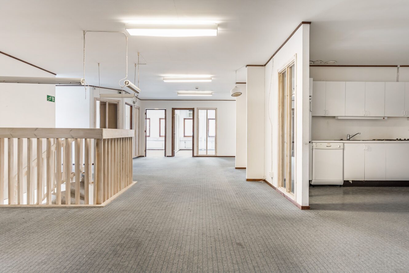 Rymlig, tom kontorslokal med heltäckningsmatta, pentry till höger och träräcke runt en trappa till vänster. Lysrörsbelysning ovanför taket.