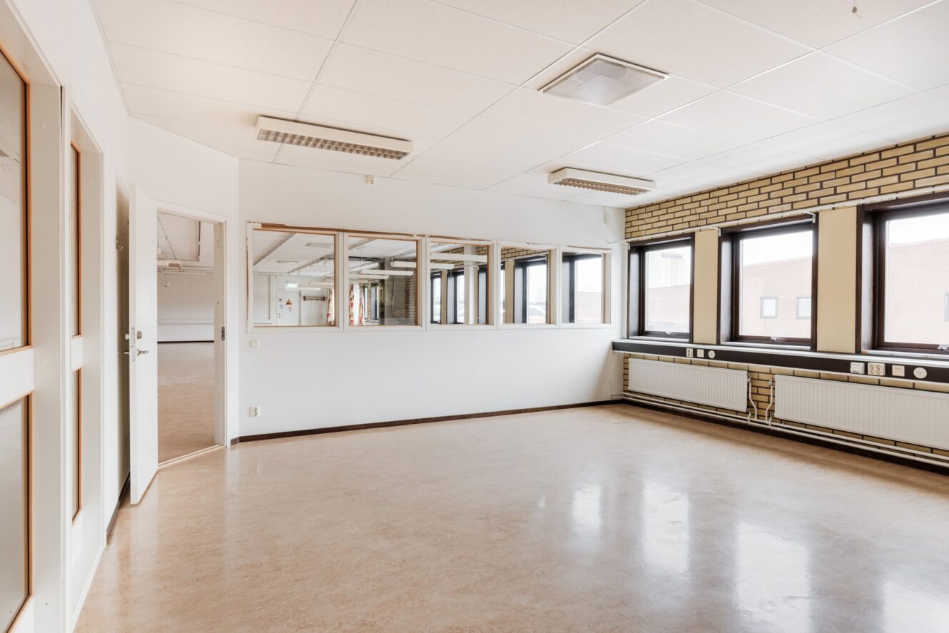 Tomt, väl upplyst rum med stora fönster, beige golv, vita takplattor, tegelvägg, väggradiator och en rad innerfönster som vetter mot ett annat utrymme.