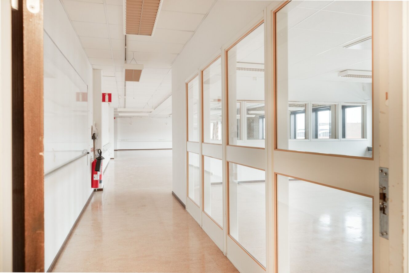 En korridor i en ljus, tom byggnad med stora innerfönster och en brandsläckare monterad på väggen.