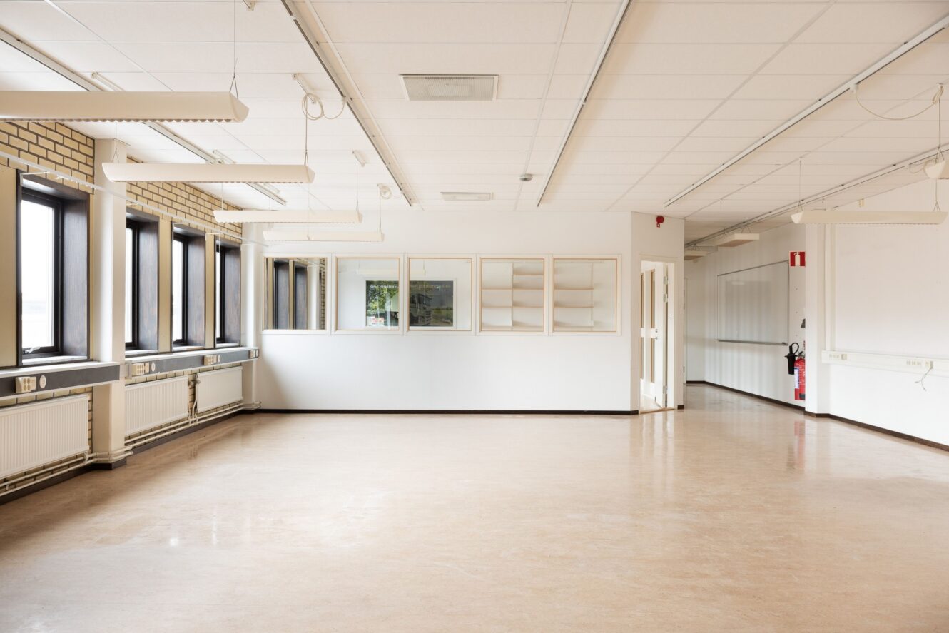 Ett tomt, väl upplyst rum med stora fönster, beige golv, vita väggar, taklampor och en brandsläckare monterad nära utgången.