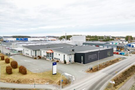 En stor industribyggnad med en svart sektion märkt PEN är omgiven av parkeringsplatser, vägar och andra kommersiella strukturer i ett industriområde.