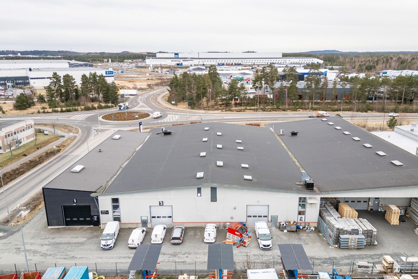 Flygfoto över ett industriområde med lagerbyggnader, parkerade skåpbilar, byggmaterial, en rondell och omgivande träd under en mulen himmel.