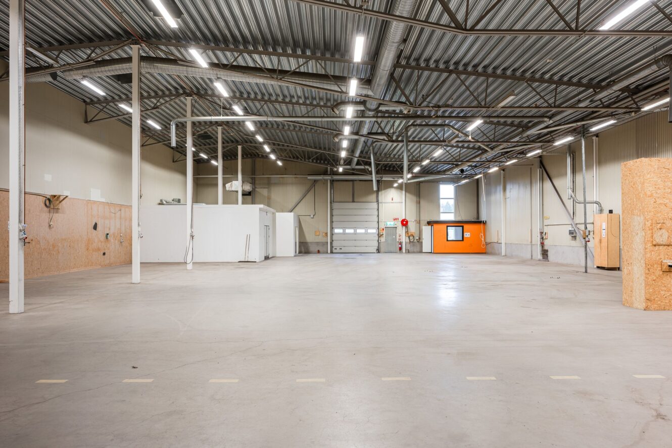 Rymlig, tom industrilokal med högt i tak, synliga bjälkar, flera taklampor, ett litet orange kontor och en stor garageport.
