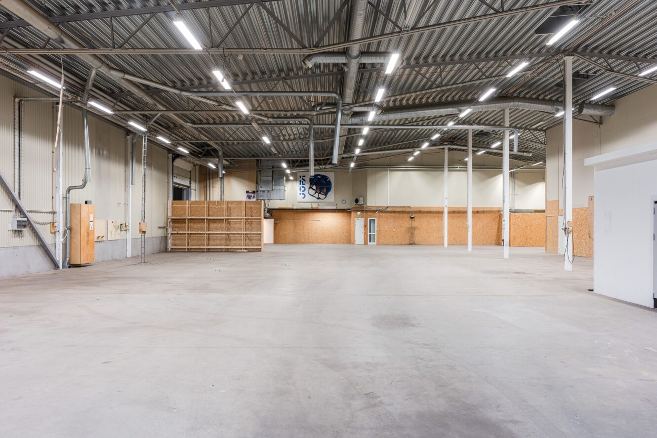Stort, tomt industrilager med högt i tak, synliga bjälkar, betonggolv, lysrörsbelysning och några partiella träväggar längs bakväggen.