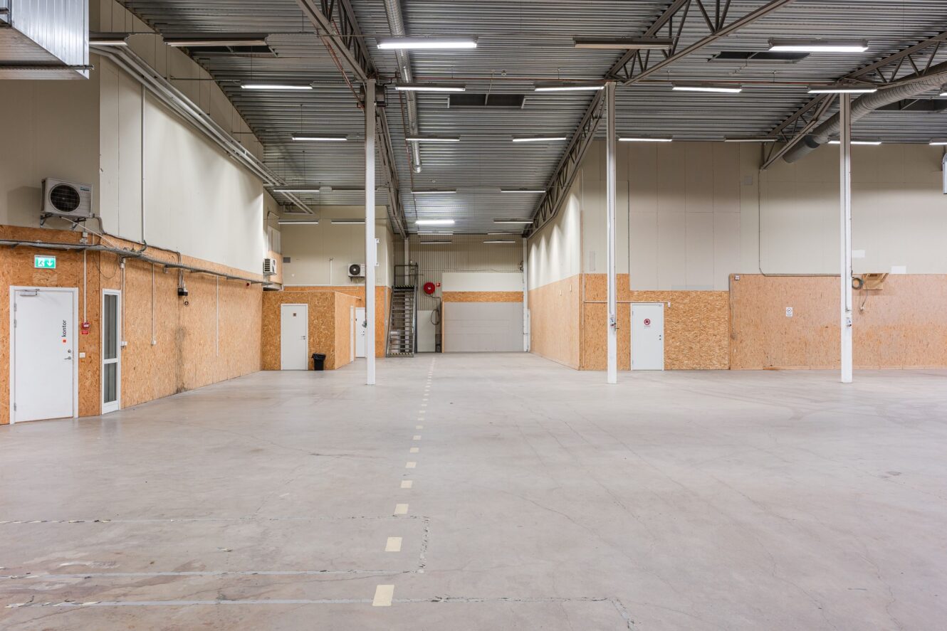 Stor tom lagerlokal med betonggolv, högt i tak, synliga bjälkar, väggar med träpanel och flera dörrar längs sidorna. Lysrör hänger från taket.