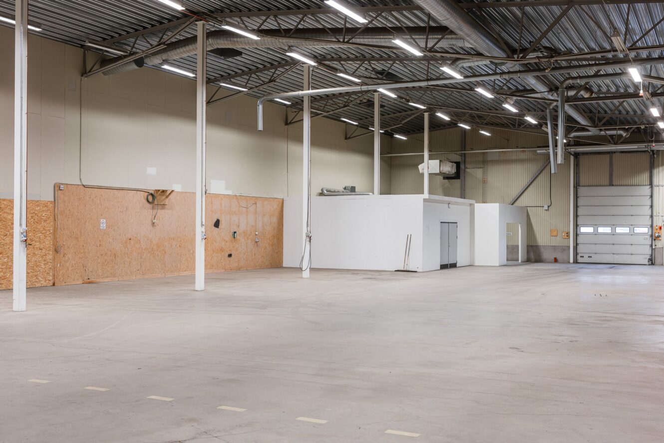 Stor tom industrilagerlokal med högt i tak, synliga rör, vita rumsindelningar, betonggolv och en stängd takskjutport.