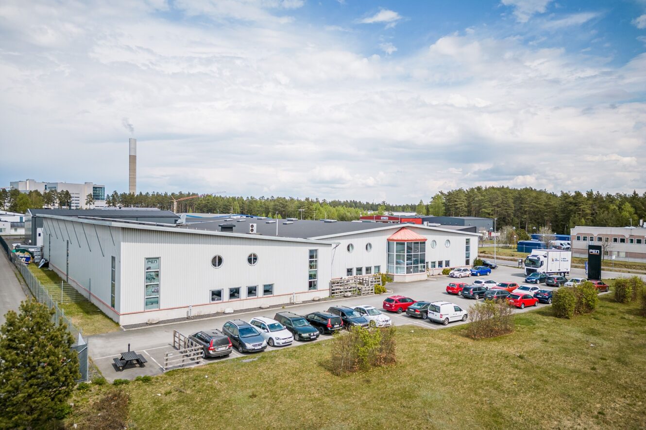 En stor industribyggnad med en parkeringsplats full av bilar framför, omgiven av gräs och träd, under en delvis molnig himmel.