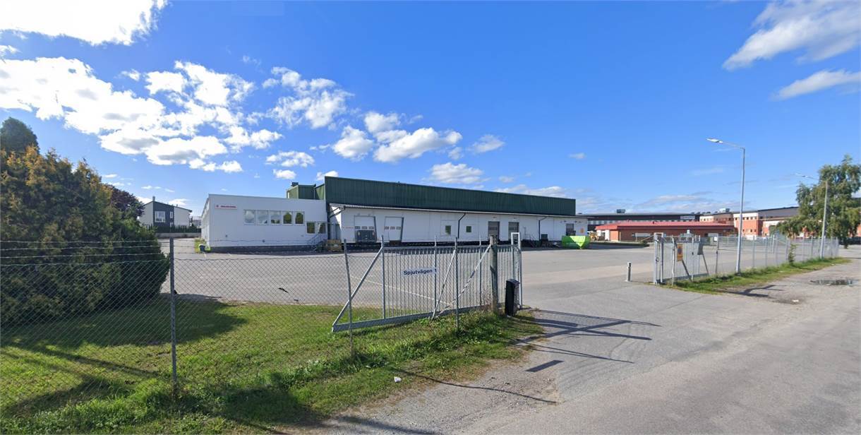 En inhägnad industribyggnad med grönt tak, lastkajer och en asfalterad tomt under en delvis molnig himmel.