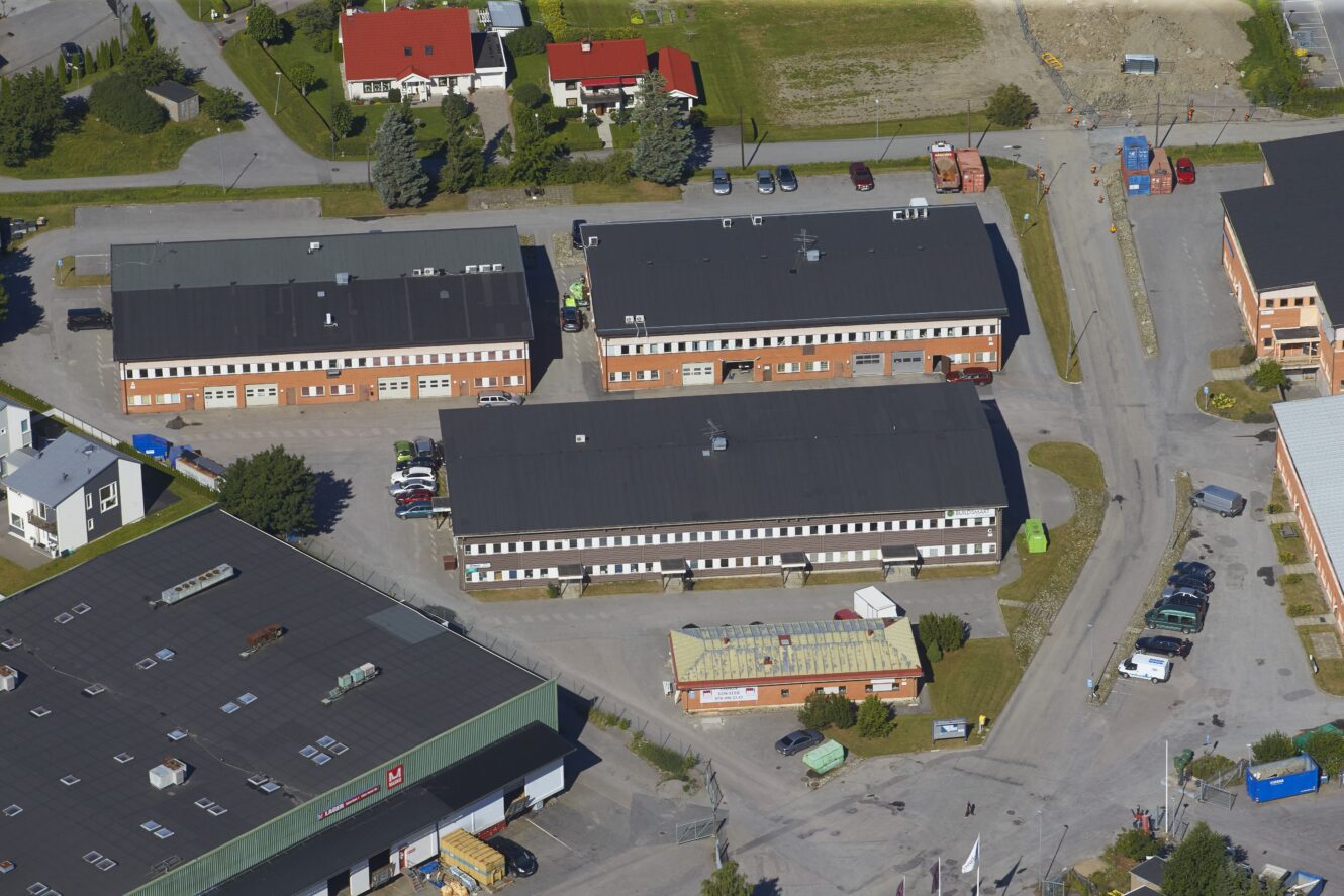 Flygfoto över ett litet industriområde med flera rektangulära lagerbyggnader, en parkeringsplats med flera fordon och närliggande bostadshus.