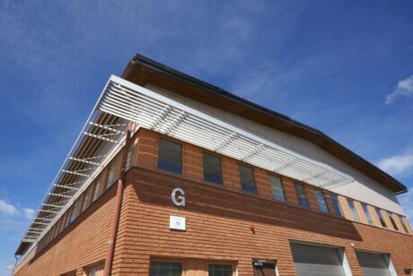 En tvåvånings tegelbyggnad med ett vitt horisontellt lamellformat solskydd på övervåningen och en stor bokstav G på ytterväggen, under en klarblå himmel.