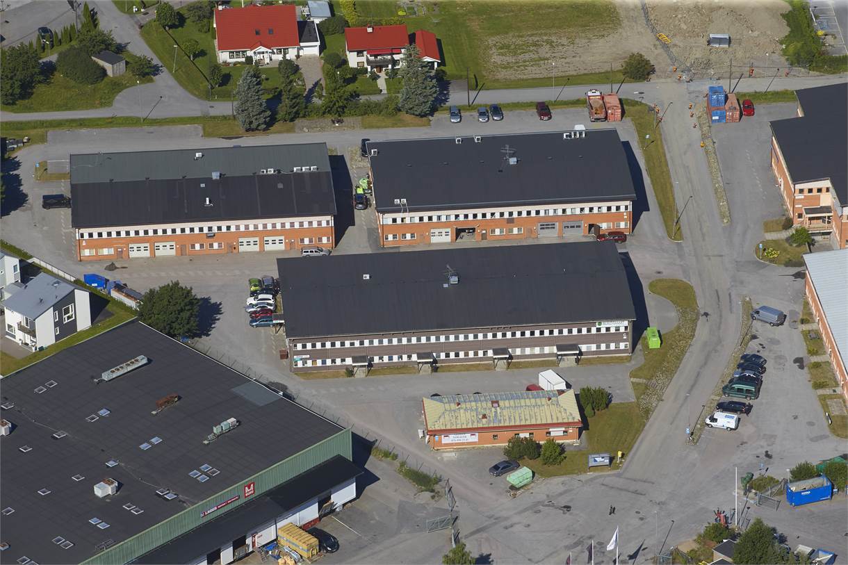 Flygfoto över ett industriområde med flera stora rektangulära byggnader, parkerade bilar och omgivande vägar. Bostadshus syns i bakgrunden.