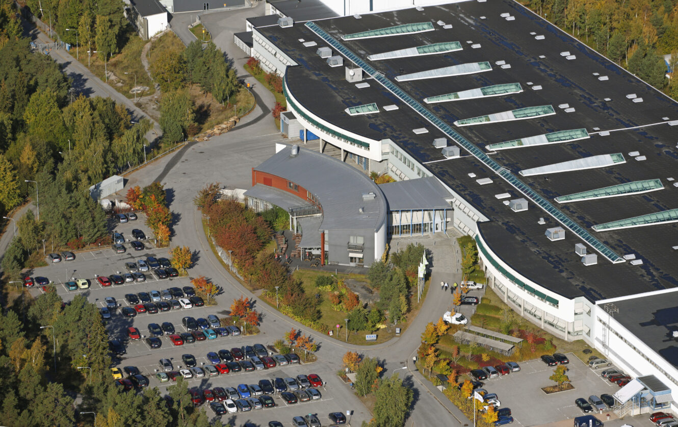 Flygfoto över en stor industri- eller forskningsanläggning med en svängd huvudentré, en intilliggande parkeringsplats fylld med bilar och omgivande träd som visar tidiga höstfärger.