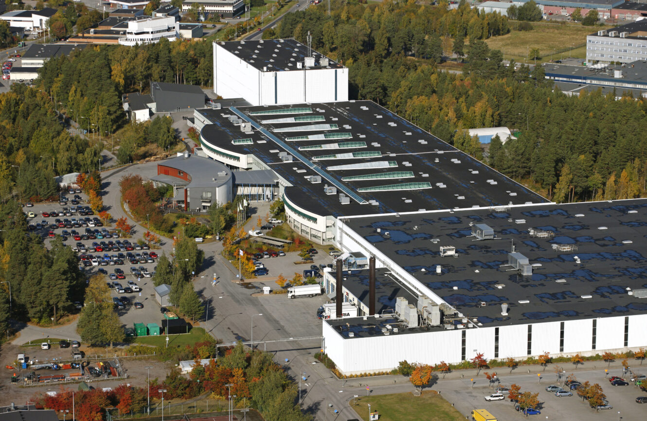 Flygfoto över en stor industrianläggning med flera byggnader, parkeringsplatser och omgivande träd i ett höstlandskap.