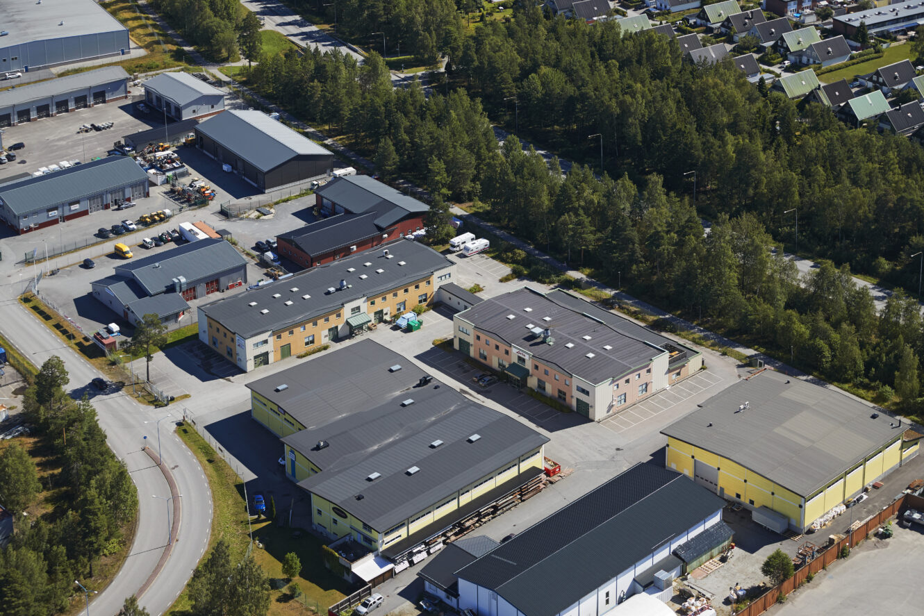 Flygfoto över en industripark med flera lager- och kontorsbyggnader omgivna av träd och ett intilliggande bostadsområde.
