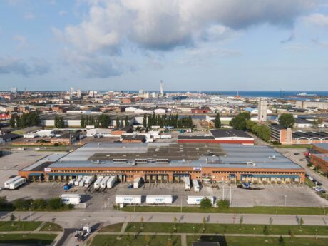 Flygfoto över en stor lagerlokal med flera lastkajer och lastbilar parkerade utanför, belägen i ett industriområde nära ett vattendrag.