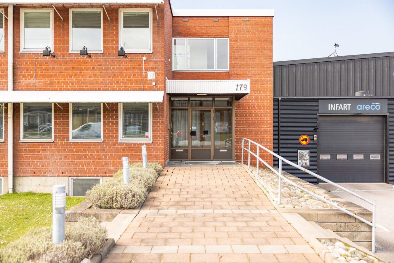 Kommersiell byggnad i rött tegel med glasdörrar vid entrén, märkt 179. Intill ligger en svart byggnad med en garageport märkt INFART och areco. Asfalterad gångväg i förgrunden.