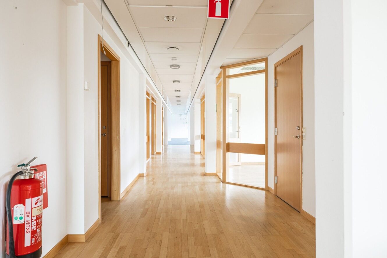En ren, tom hall med ljusa trägolv, vita väggar, trädörrar och en brandsläckare monterad på den vänstra väggen.