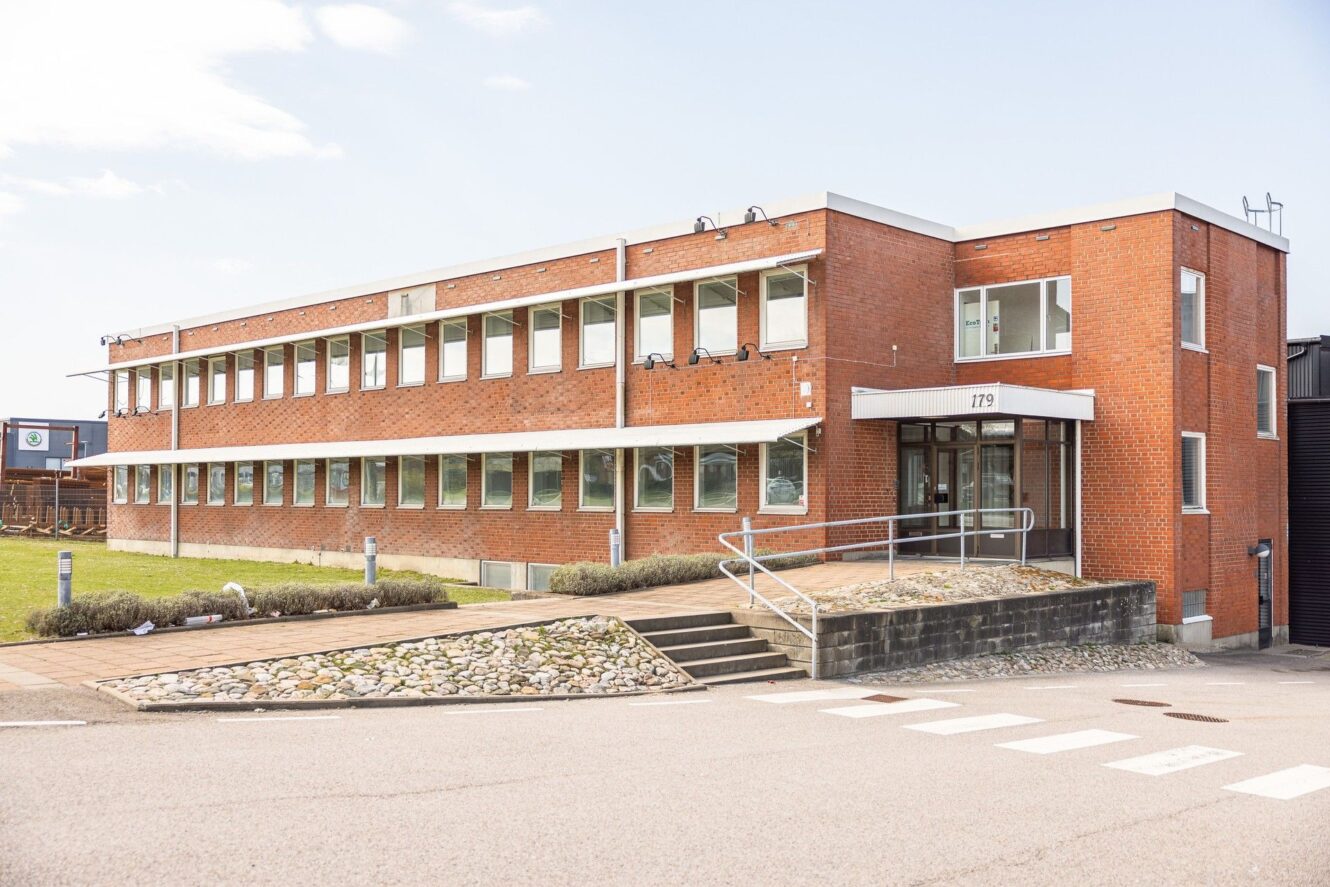 Kontorsbyggnad i tegel i två våningar med stora fönster, ramp och trappa vid entrén, omgiven av en asfalterad yta och viss landskapsarkitektur.