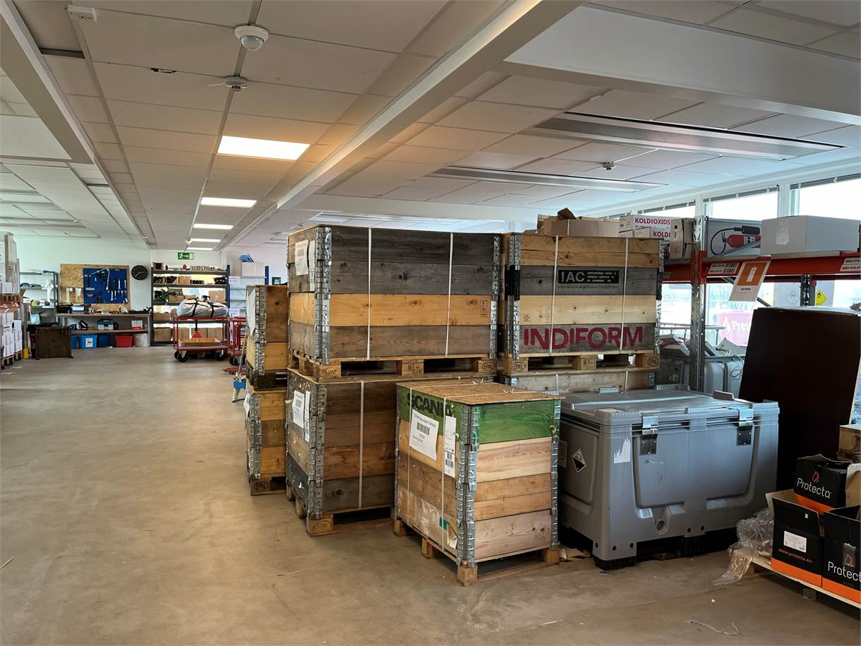 Stora trälådor och industriutrustning staplas på pallar i en rymlig lagerlokal med betonggolv och lysrörsbelysning.