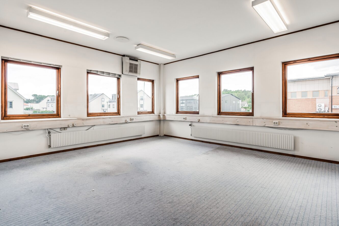 Tomt, omöblerat rum med vita väggar, flera fönster, lysrör i taket och radiatorer under fönstren. Byggnader är synliga utanför.