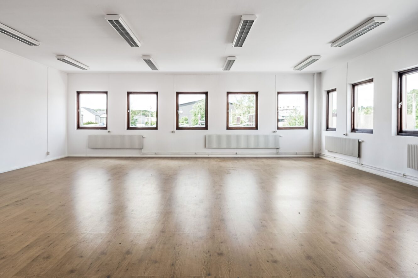Ett tomt, väl upplyst rum med vita väggar, trägolv, sex fönster och flera taklampor.
