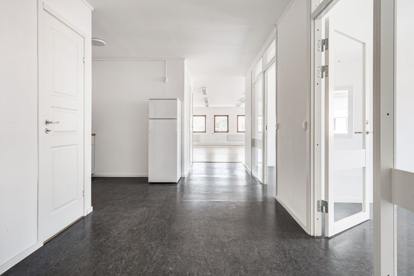 En ljus, tom kontorskorridor med vita väggar, svarta golv, flera öppna dörrar och ett vitt kylskåp mot väggen.