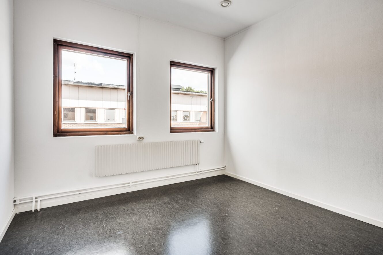Litet, tomt rum med två fönster, vita väggar, mörkt golv och en radiator under fönstren. Naturligt ljus kommer in från utsidan.