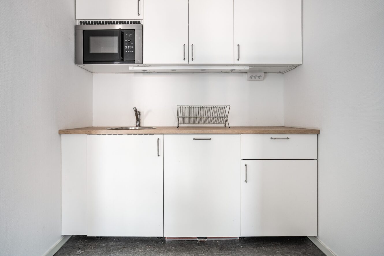 Ett litet, minimalistiskt pentry med vita skåp, en bänkskiva i trä, en diskho, ett diskställ och en svart mikrovågsugn monterad ovanför.