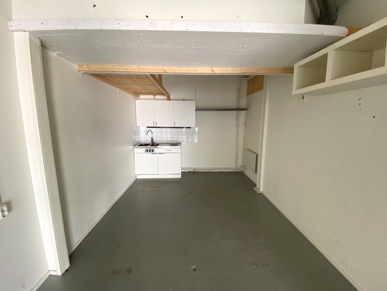 Litet, omöblerat rum med pentry, vita skåp, diskho och spis. Utrymmet har ett grått golv, vita väggar och ett loft i trä ovanför köket.
