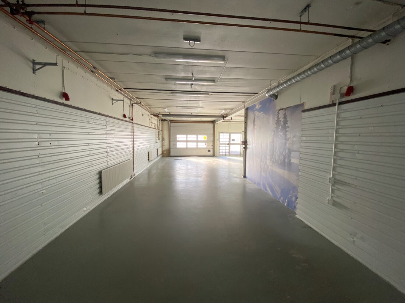 Ett tomt, industriellt inomhusutrymme med vita väggar, rörledningar, ett betonggolv och en stor garageport längst bort.