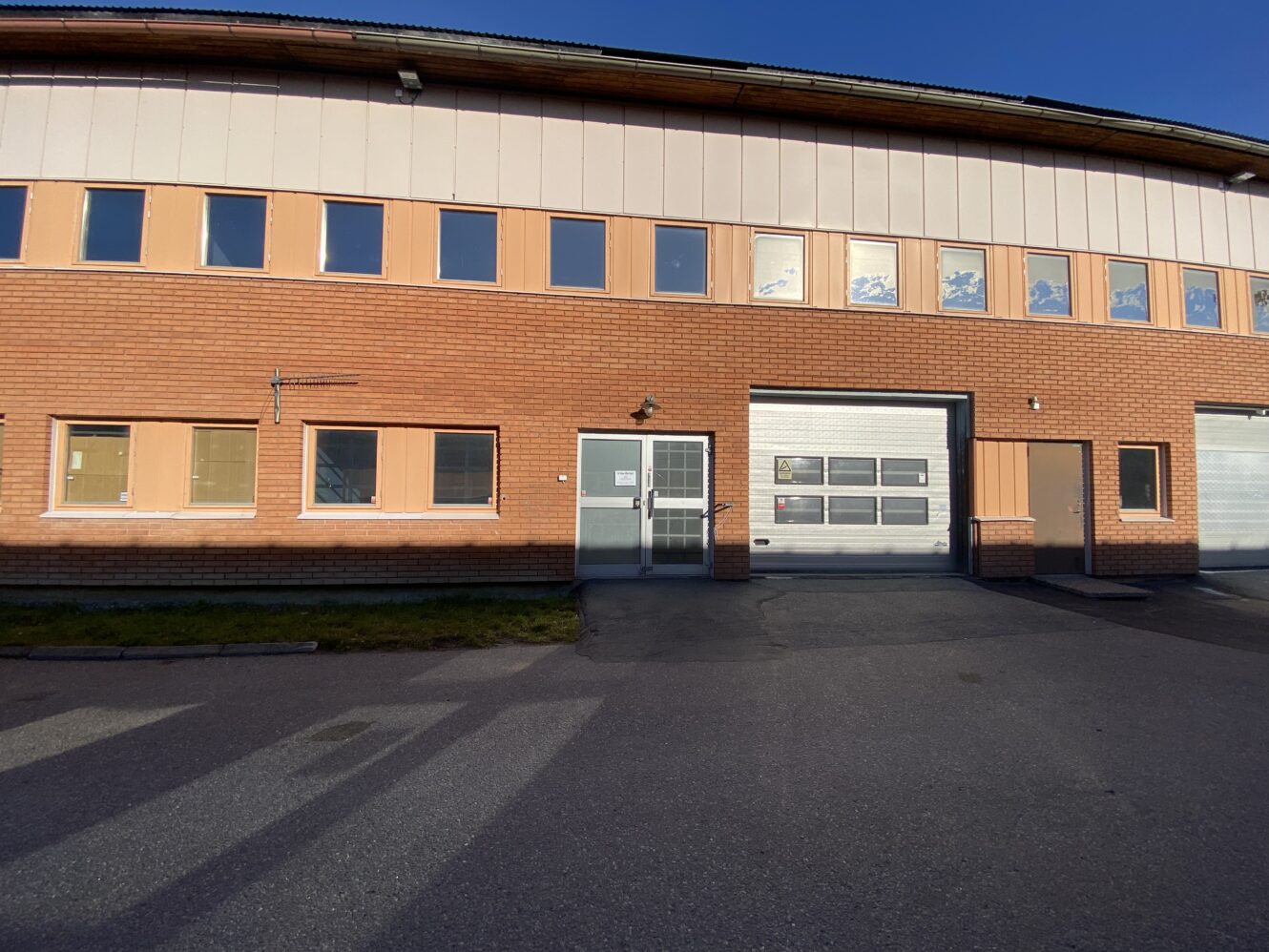 En industribyggnad i tegel och beige med rader av fönster, en glasdörr och två stora garageportar, sedd från en asfalterad parkeringsplats.