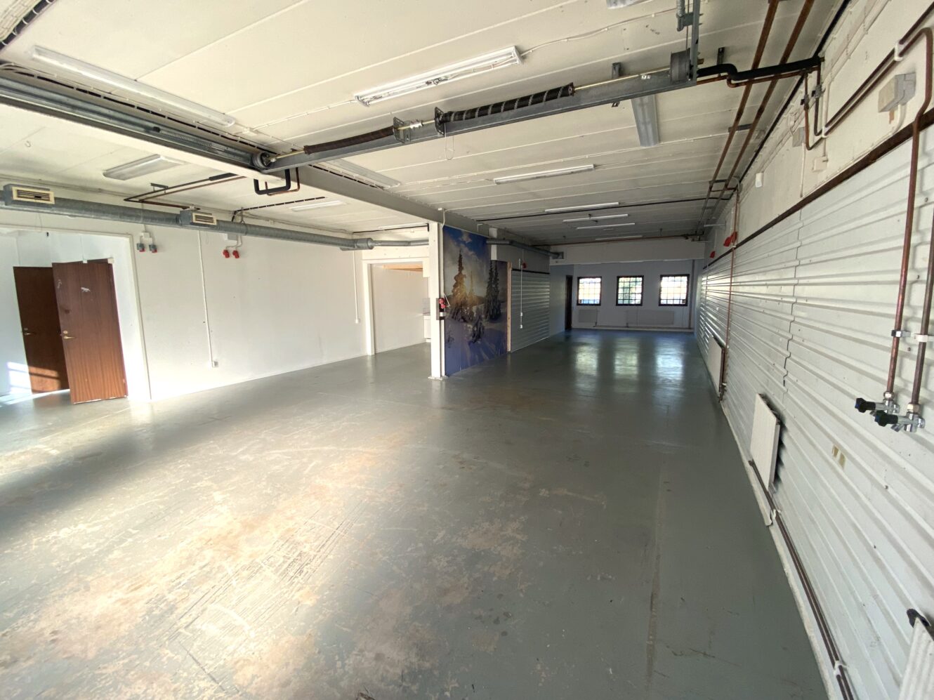 Ett tomt rum i industriell stil med betonggolv, väggar med vit panel, synliga rör och lysrörsbelysning. Tre fönster och en väggmålning syns i bortre änden.