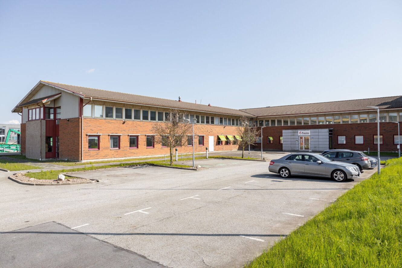 En modern tegelbyggnad i två våningar med stora fönster, en liten parkeringsplats med tre bilar och grönt gräs i förgrunden under en klar himmel.