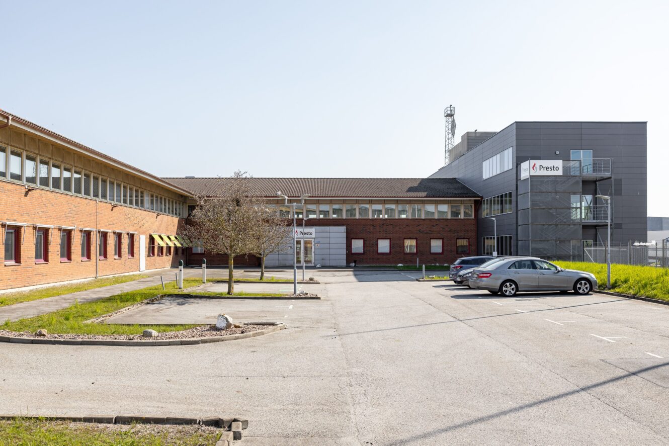 En modern kontorsbyggnad med tegel och grå fasad, märkt Presto, har en nästan tom parkeringsplats med två parkerade bilar och klart, soligt väder.