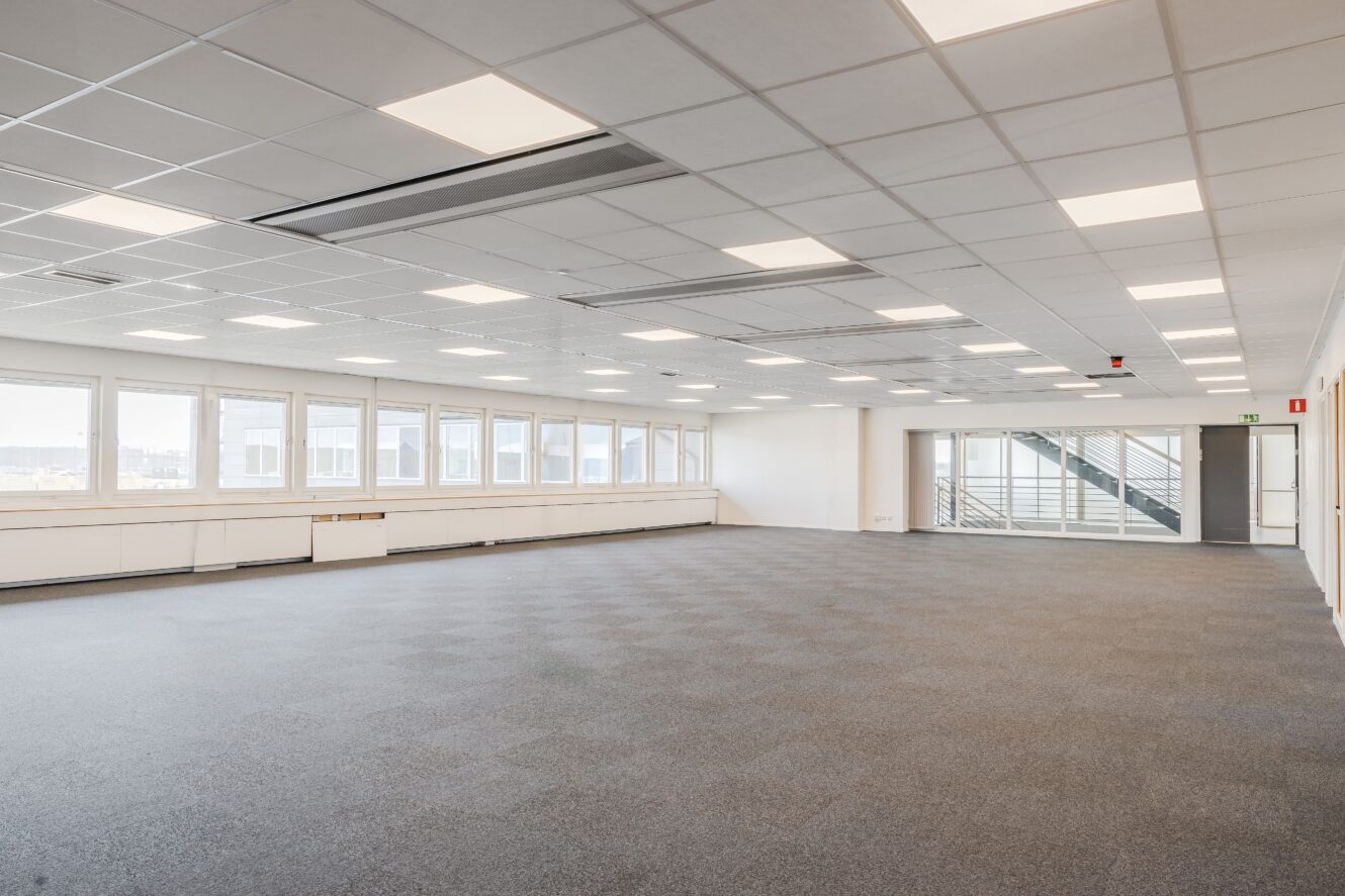 Stort tomt kontorsutrymme med heltäckningsmatta, vita väggar, ett gallertak med lysrör, stora fönster och en trappa i bakgrunden.